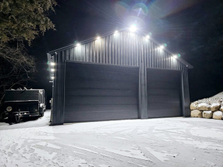 A well-lit black pole barn company shop featuring seamless gutters installed by The Gutter Guys, with snow-covered ground and a work trailer nearby