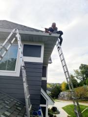 Man installing seamless gutters on a home in Minnesota, with gorgeous landscaping in the background