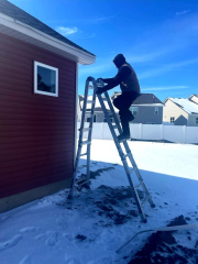 A Call The Gutter Guys professional climbing a ladder to install or maintain seamless gutters on a red house during winter. The snowy background showcases a residential neighborhood with clear blue skies