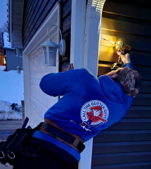 A professional from Call The Gutter Guys installing a seamless gutter at a home's exterior during winter, using a drill while wearing a blue company hoodie