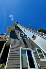 A gutter installation expert from Call The Gutter Guys working at a high elevation on a tall ladder against a multi-story house under a clear blue sky